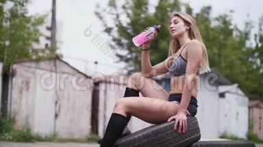 一位年轻漂亮的运动型金发女郎穿着上衣和短裤，坐在轮胎上，喝着瓶子里的水